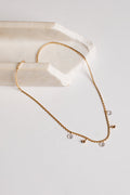 A delicate gold chain necklace with small clear crystal pendants and tiny gold beads, displayed on a white surface next to a cream-colored stone prop.