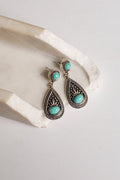 A pair of silver-toned teardrop dangle earrings with oval turquoise stones, displayed on a textured white stone prop against a light neutral background.