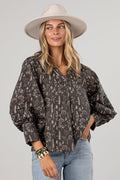 A woman wearing a dark brown and beige floral print blouse with voluminous balloon sleeves, paired with light blue jeans and a beige wide-brim hat, against a solid light gray background.