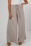 A person wearing wide-leg pleated pants in a taupe-beige color with thin vertical stripes, paired with a white top and brown sandals, shown from the waist down against a light gray studio background.